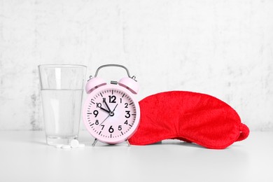 Photo of Red sleep mask, glass of water, pills and alarm clock on white table