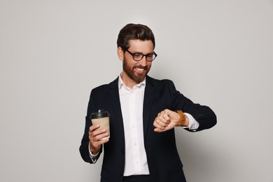Handsome bearded man with glasses and paper cup looking at wristwatch on light grey background