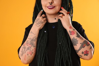 Young woman with tattoos on arms, nose piercing and dreadlocks against yellow background, closeup