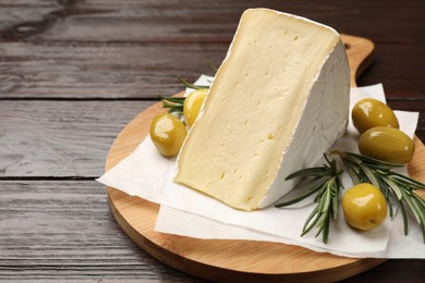 Photo of Board with piece of tasty camembert cheese, olives and rosemary on wooden table, closeup. Space for text