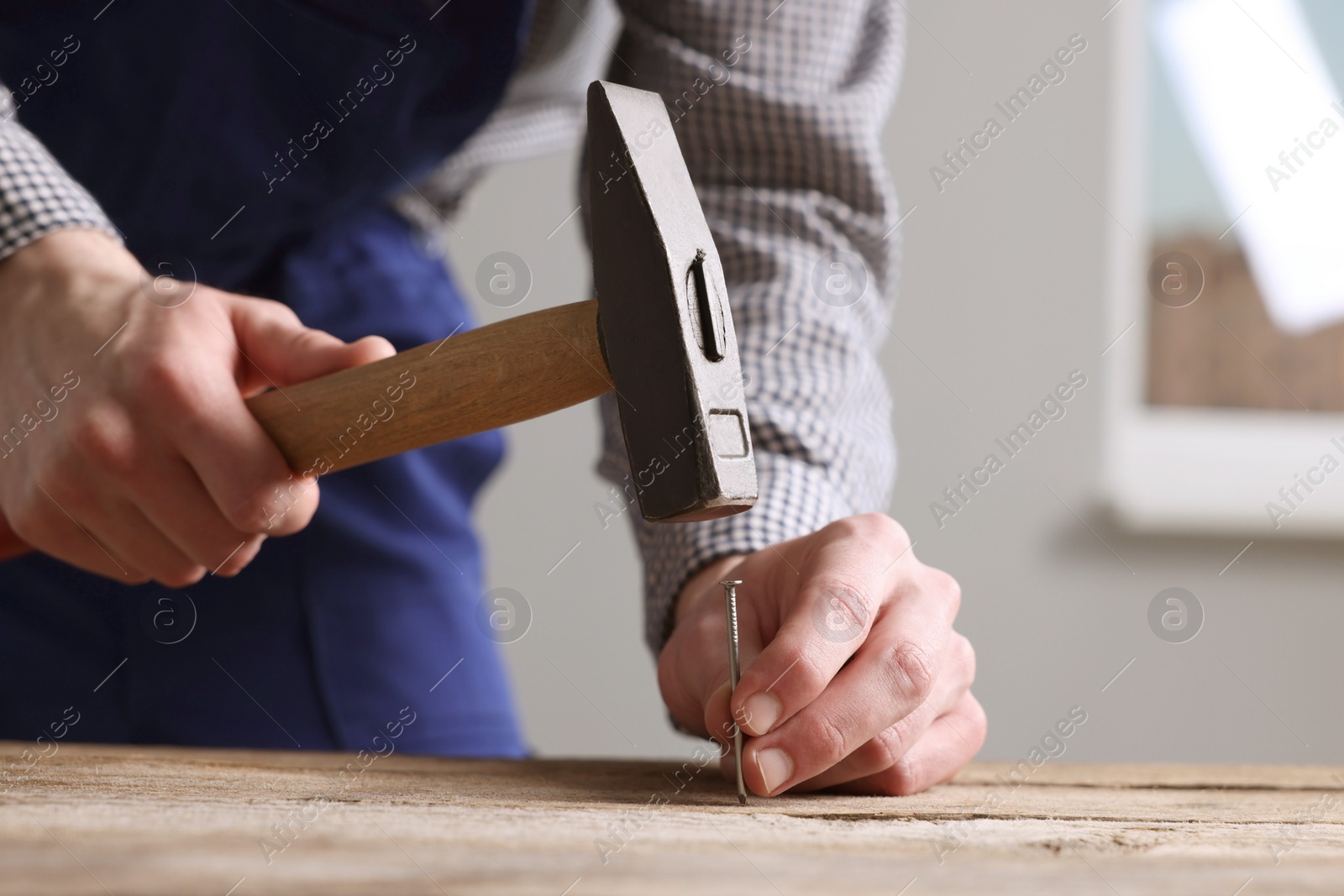 Photo of Professional repairman hammering nail into wooden board indoors, closeup