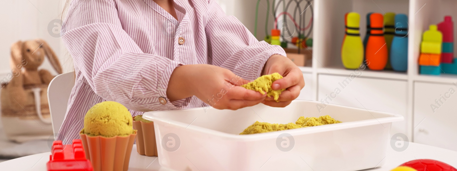 Image of Cute little girl playing with bright kinetic sand at table in room, closeup. Banner design