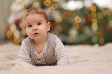 Cute little baby on knitted blanket against blurred festive lights, space for text. Winter holiday