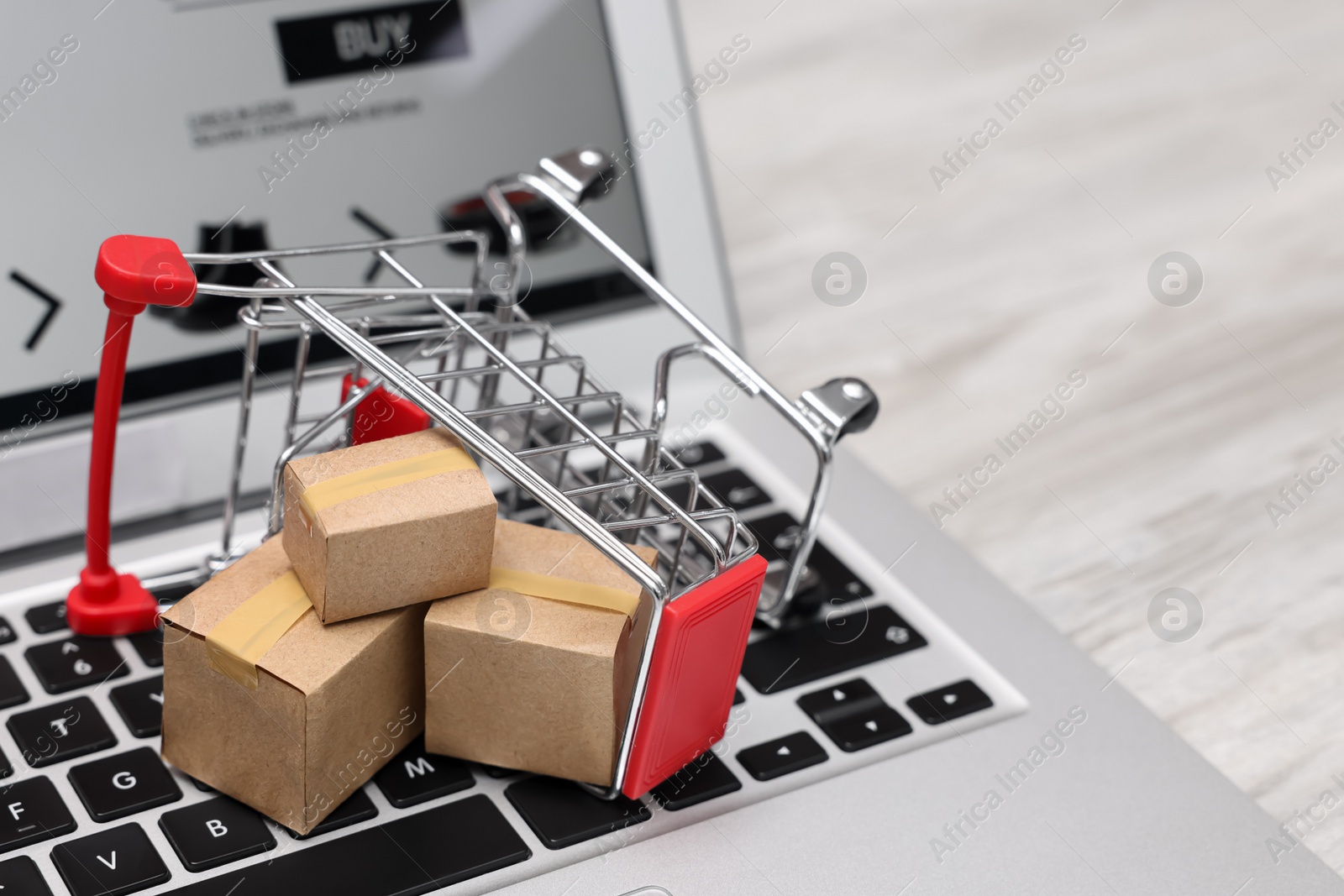 Photo of Internet store. Small cardboard boxes, shopping cart and laptop on light wooden table, closeup. Space for text