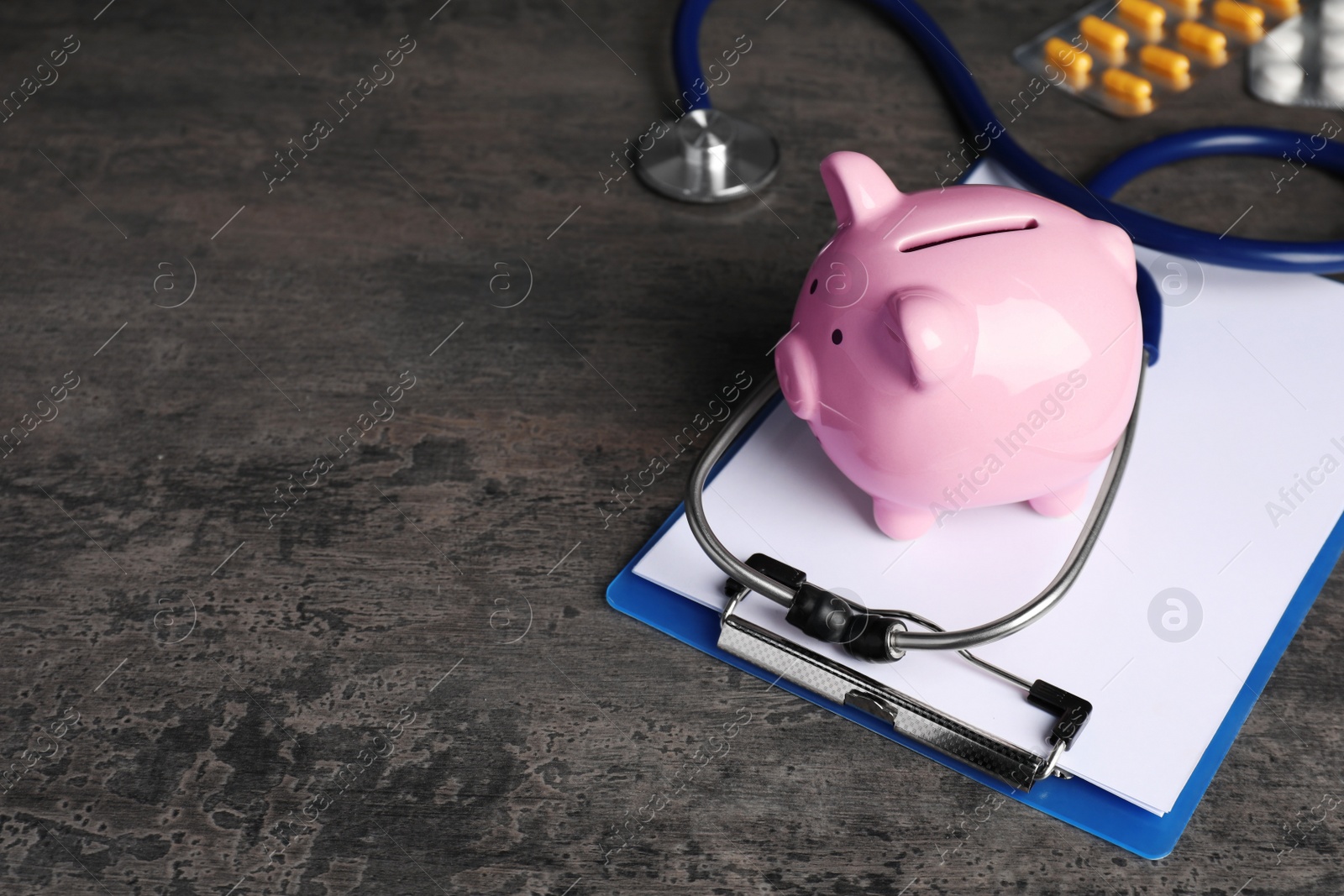 Photo of Piggy bank, clipboard, stethoscope and pills on dark grey textured table, space for text. Medical insurance