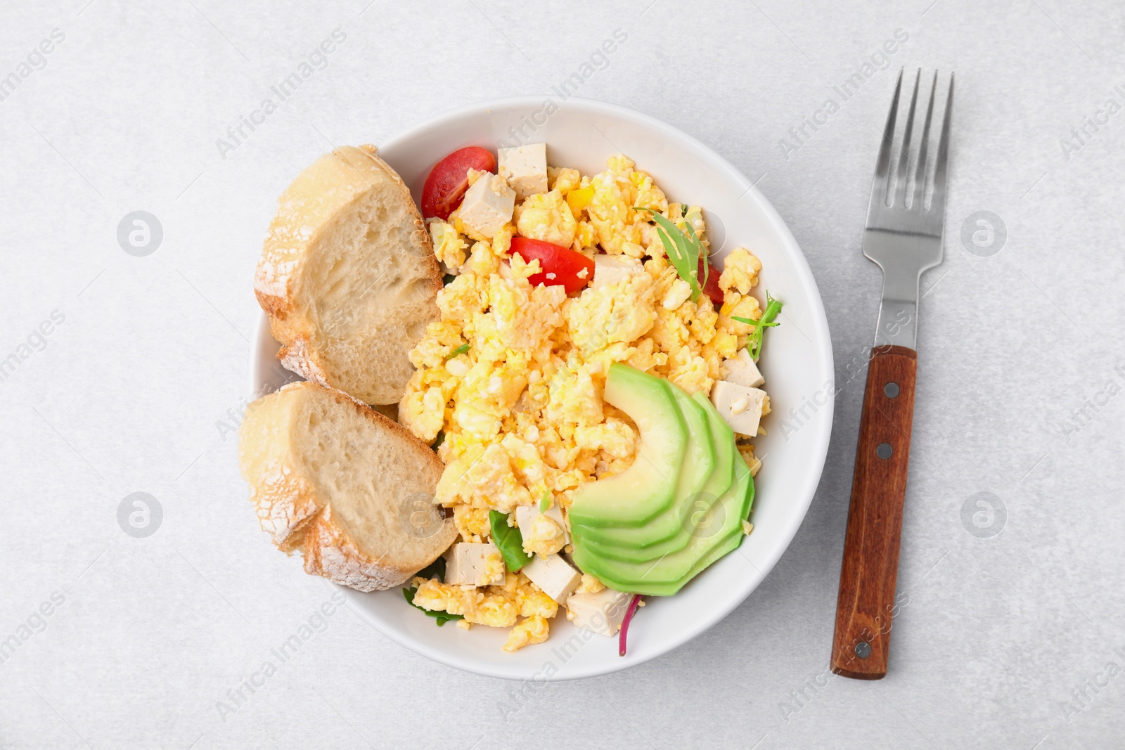 Photo of Bowl with delicious scrambled eggs, tofu, avocado and slices of baguette on white table, flat lay