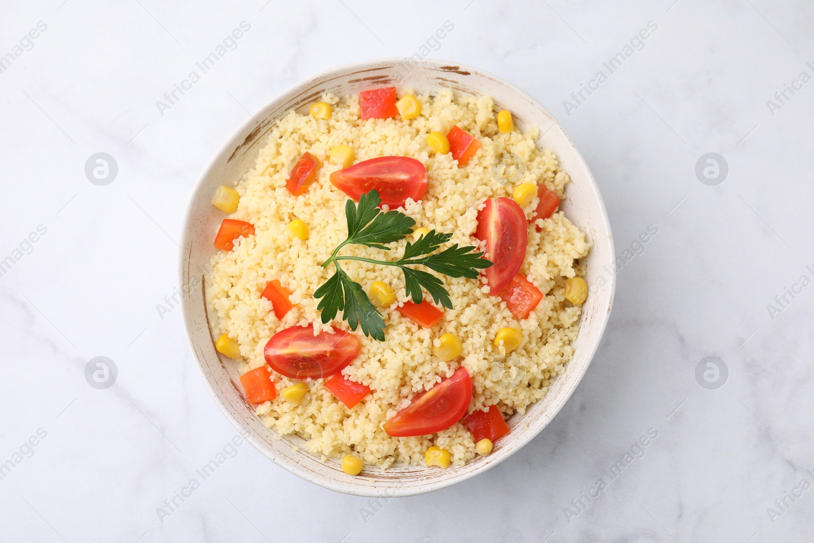 Photo of Tasty couscous with parsley, corn and tomatoes in bowl on white marble table, top view
