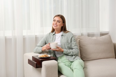 Happy woman holding cup of drink on sofa armrest wooden table at home