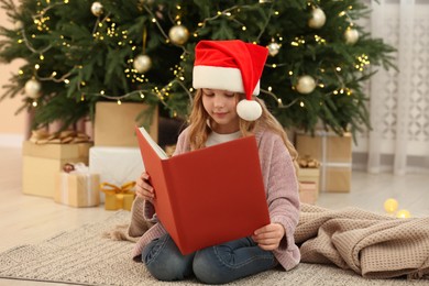 Cute little girl in Santa hat reading book near tree and gifts at home. Christmas atmosphere