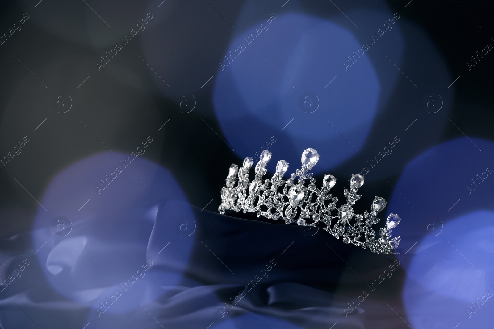 Photo of Beautiful silver tiara with diamonds on dark cloth against blurred lights, space for text