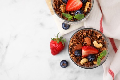 Photo of Tasty granola with berries, nuts and mint in glasses on white marble table, top view. Space for text