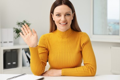 Photo of Woman waving hello during video chat at table in office, view from web camera