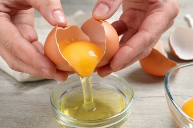 Woman separating egg yolk from white over glass bowl at wooden table, closeup