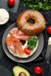 Photo of Delicious bagel with cream cheese, jamon, tomato and parsley on black table, flat lay