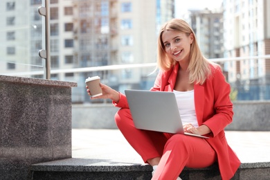 Photo of Beautiful businesswoman with laptop and cup of coffee sitting on stairs outdoors