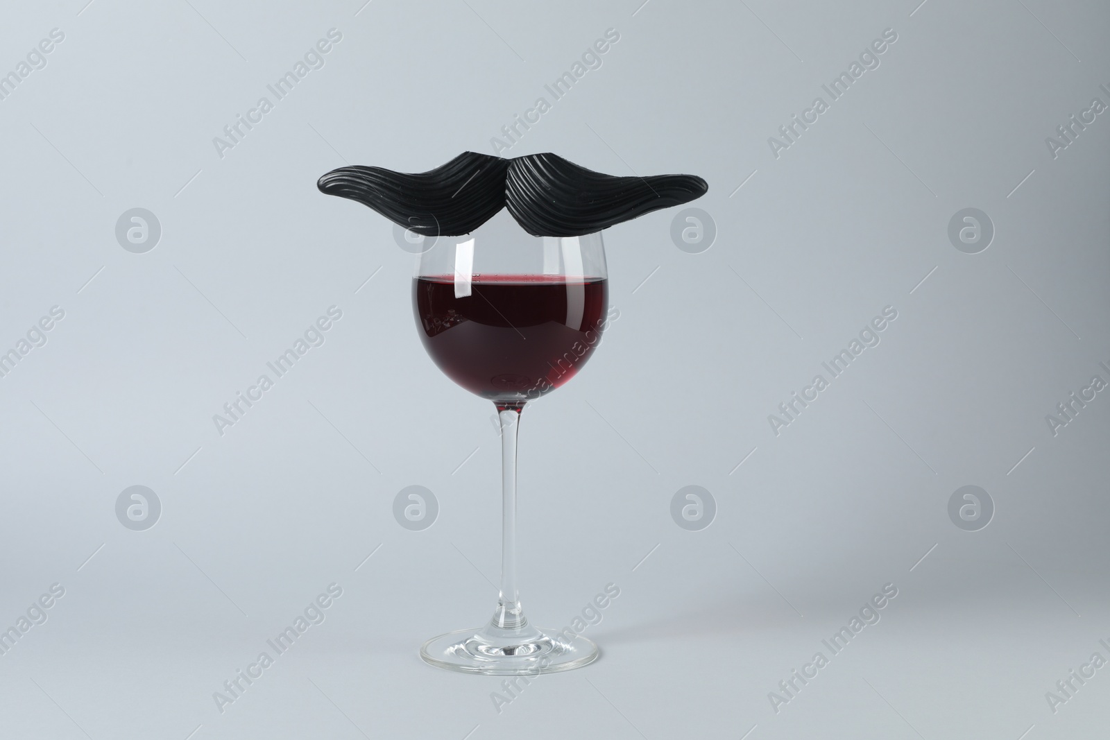 Photo of Glass of red wine with funny fake mustache on grey background