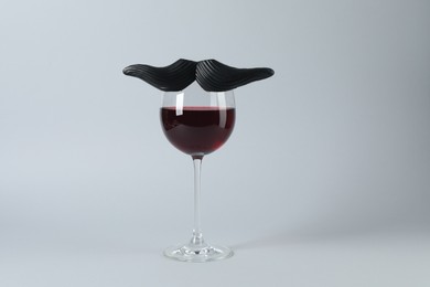 Glass of red wine with funny fake mustache on grey background