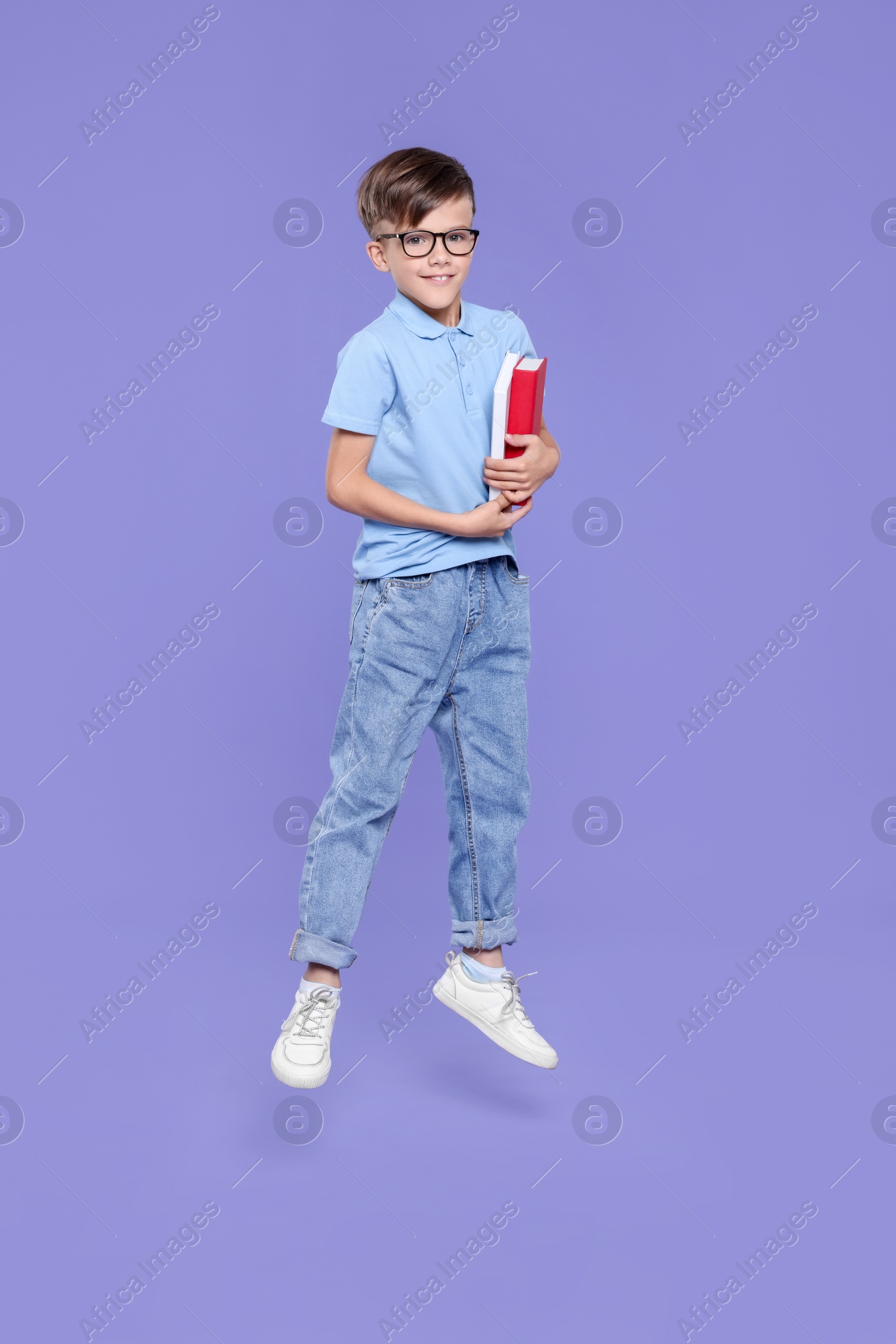 Photo of Cute schoolboy in glasses holding books and jumping on violet background