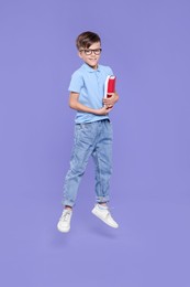 Cute schoolboy in glasses holding books and jumping on violet background