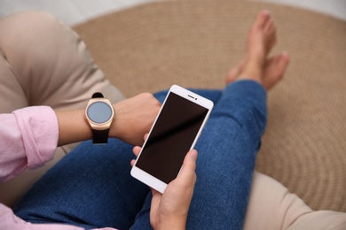 Photo of Woman with modern smartwatch and mobile phone in armchair at home, closeup
