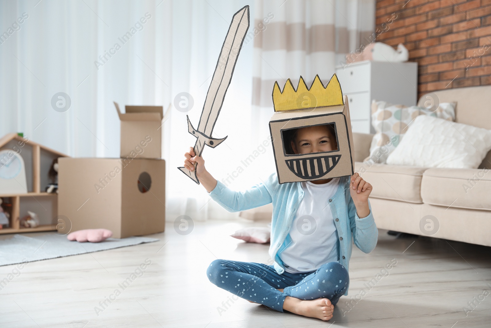 Photo of Cute little girl playing with cardboard armor in living room