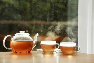 Photo of Teapot and cups of hot tea on wooden table against blurred background, space for text