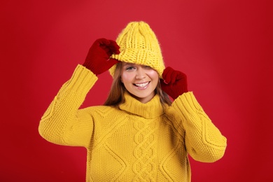 Funny young woman in warm sweater, gloves and hat on red background. Winter season