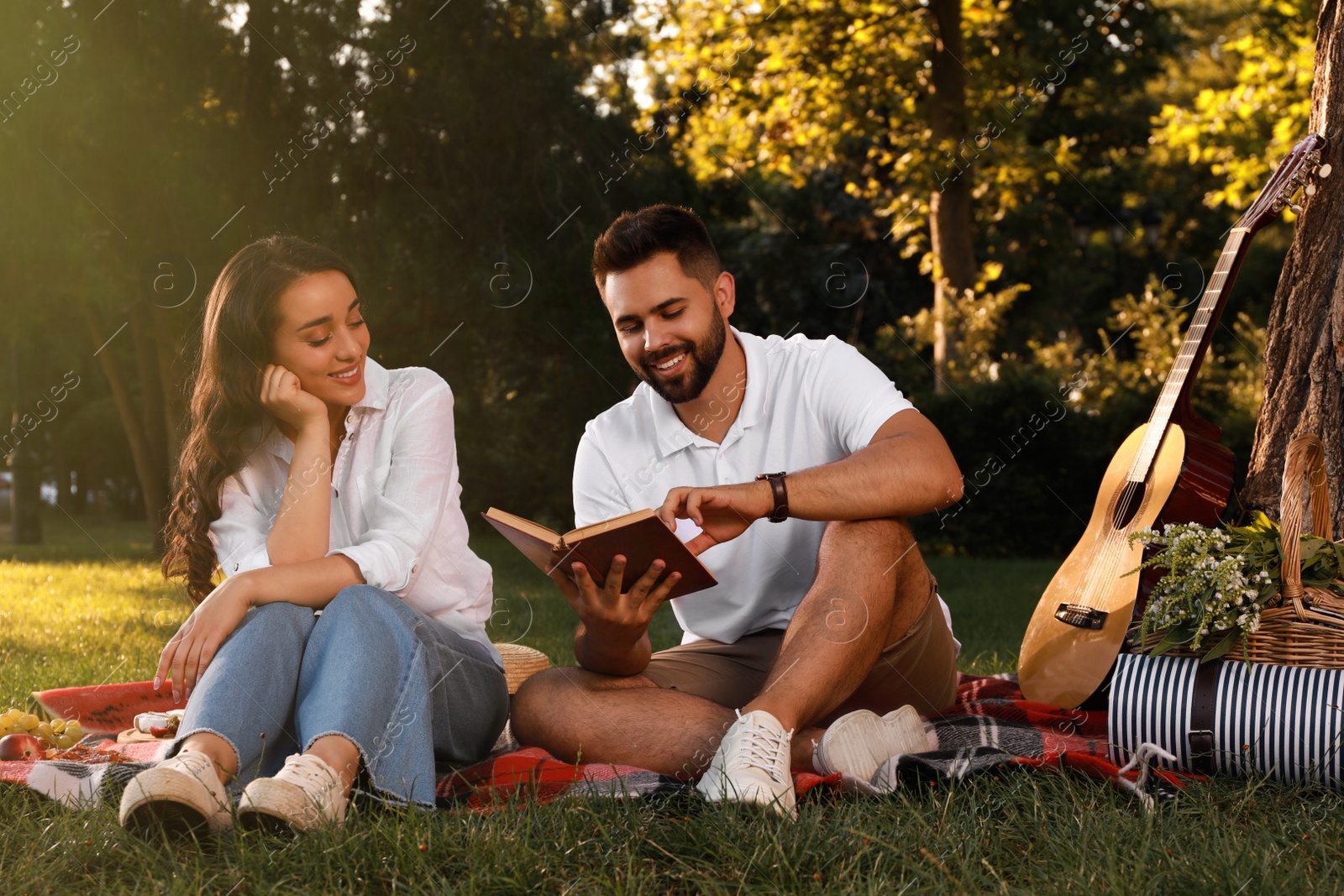 Photo of Happy young couple reading book on plaid in park. Summer picnic