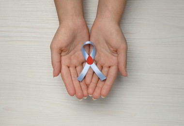 Woman holding light blue ribbon with paper blood drop at wooden table, top view. Diabetes awareness