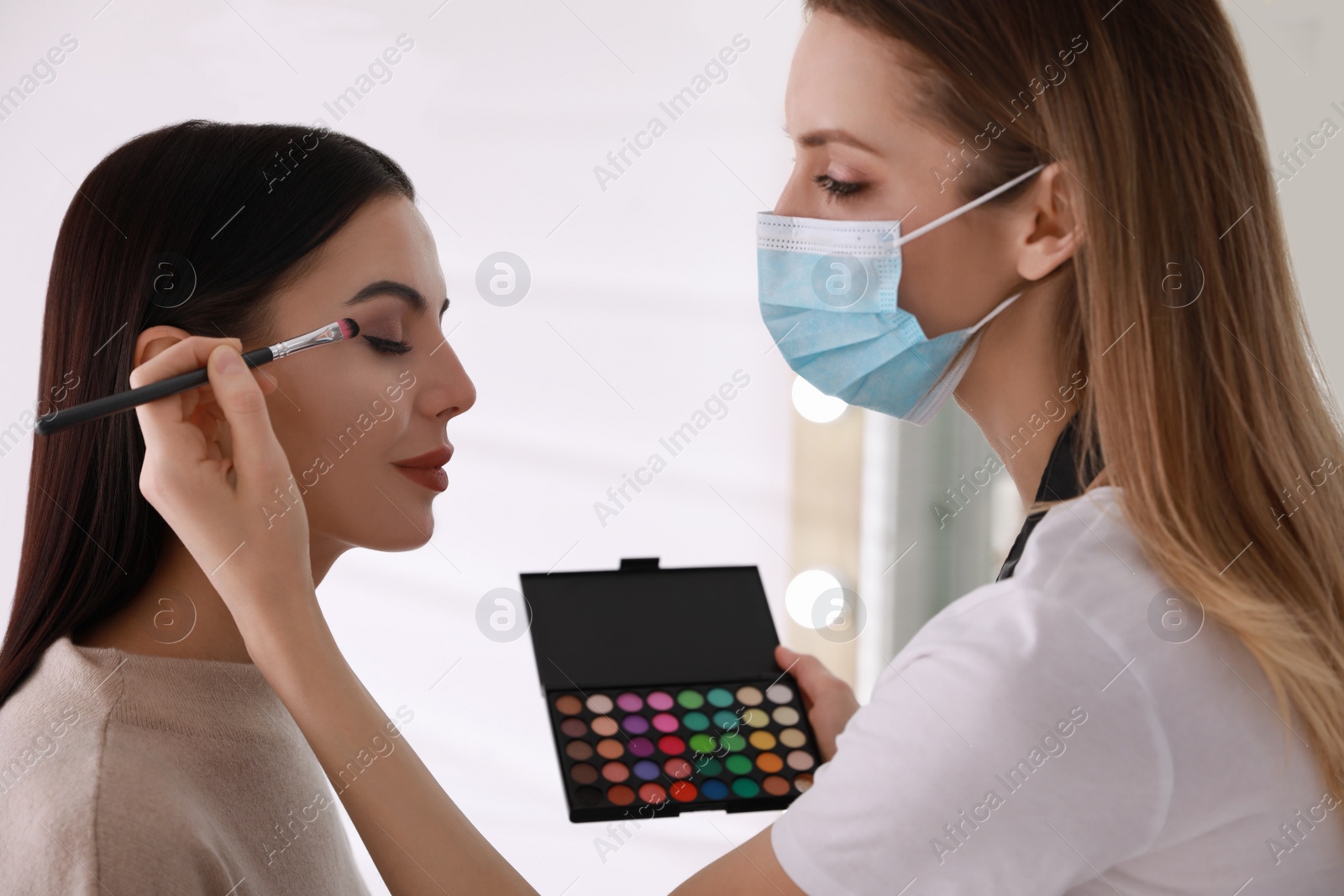 Photo of Makeup artist in protective mask working with woman indoors. Preventive measures during COVID-19 pandemic