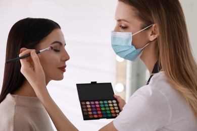 Makeup artist in protective mask working with woman indoors. Preventive measures during COVID-19 pandemic