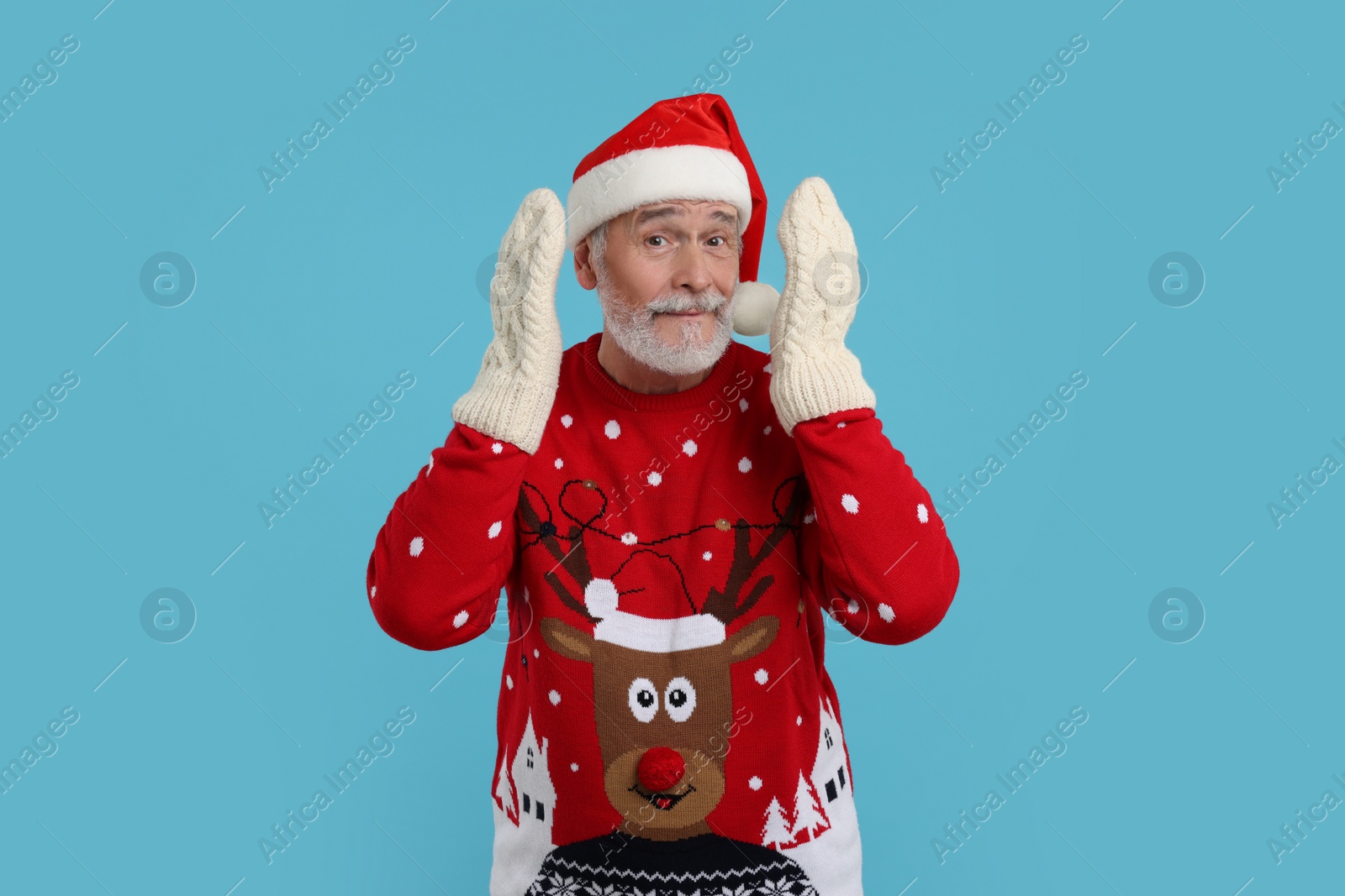 Photo of Senior man in Christmas sweater, Santa hat and knitted mittens on light blue background