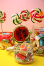 Tasty colorful candies in glass jars on yellow table against pink background