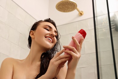 Happy woman with bottle of shampoo in shower at home, low angle view. Washing hair