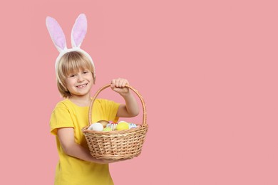 Photo of Happy boy in bunny ears headband holding wicker basket with painted Easter eggs on pink background. Space for text