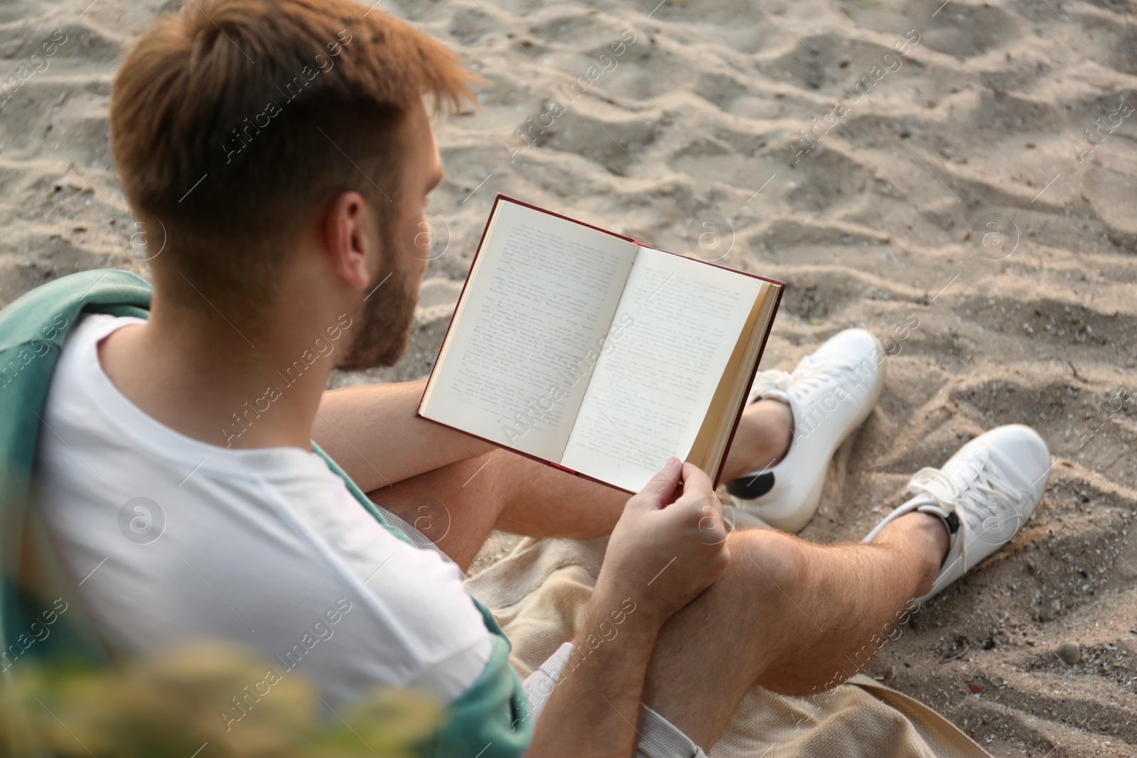 Photo of Young man reading book on sandy beach, back view