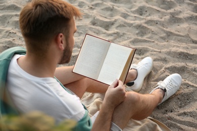 Young man reading book on sandy beach, back view