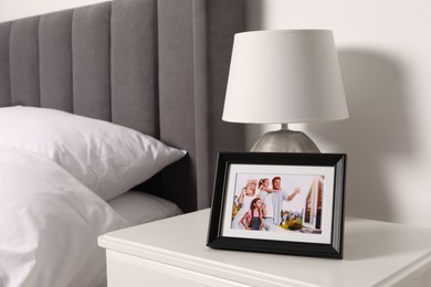 Photo of Frame with family photo and lamp on white bedside table in bedroom
