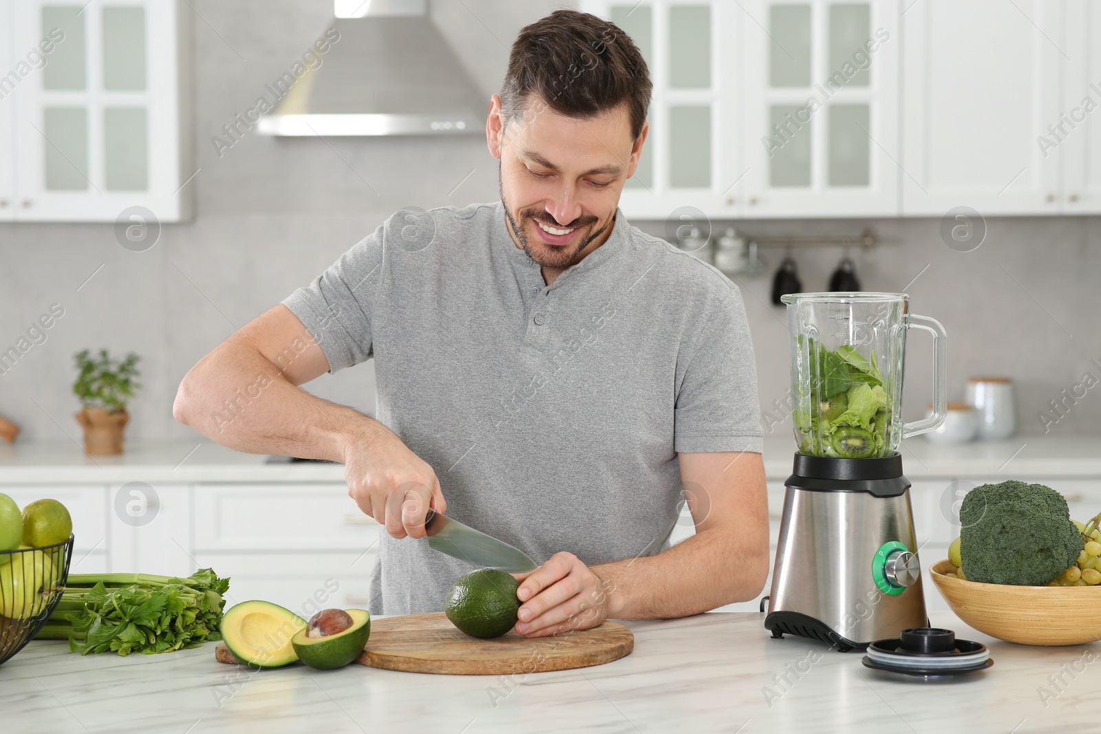 Photo of Happy man cutting avocado for delicious smoothie at white marble table in kitchen