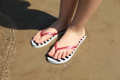 Woman in stylish flip flops on sandy beach near sea, closeup