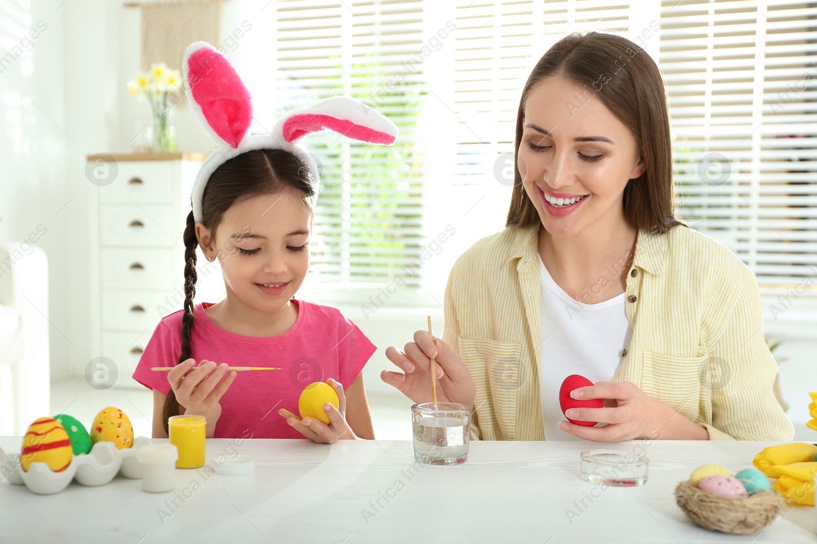 Photo of Happy daughter with bunny ears headband and her mother painting Easter eggs at home