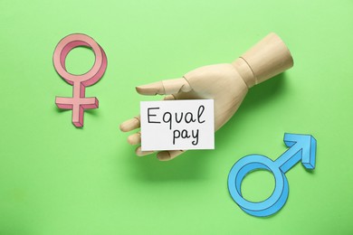 Photo of Equal pay. Wooden mannequin hand, paper note, gender symbols on green background, flat lay