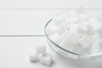 Many sugar cubes in glass bowl on white wooden table, closeup. Space for text