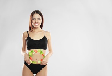 Image of Slim young woman wearing underwear and images of vegetables on her belly against light background. Healthy eating