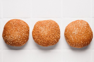 Photo of Fresh buns with sesame seeds on white tiled table, flat lay