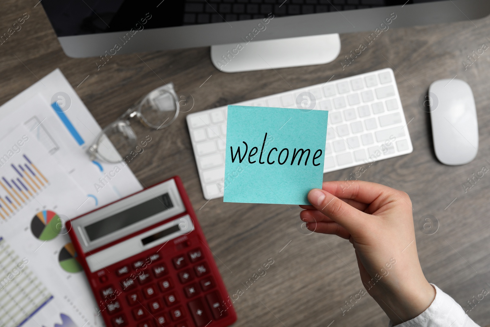 Image of Woman holding paper note with word Welcome over her office desk, top view