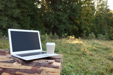 Photo of Modern laptop with blank screen and coffee cup on log in nature, space for text. Working outdoors