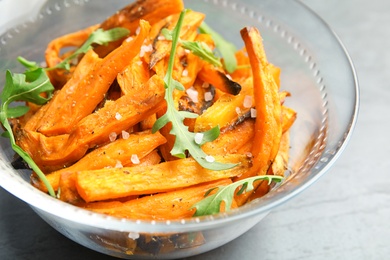 Photo of Glass bowl with baked sweet potato slices and arugula on grey table, closeup