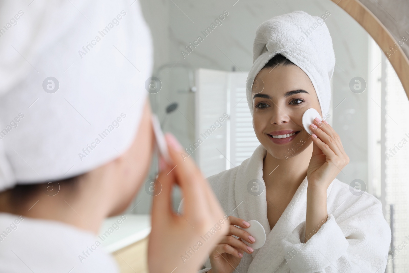 Photo of Beautiful young woman cleaning her face with cotton disk near mirror in bathroom