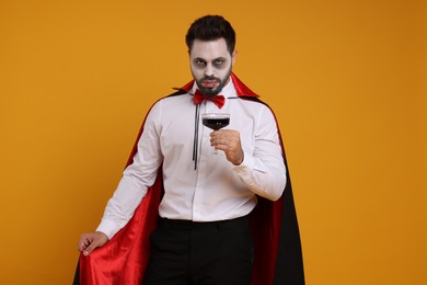 Man in scary vampire costume with fangs and glass of wine on orange background. Halloween celebration